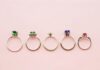Five gemstone rings arranged side by side on a pink background